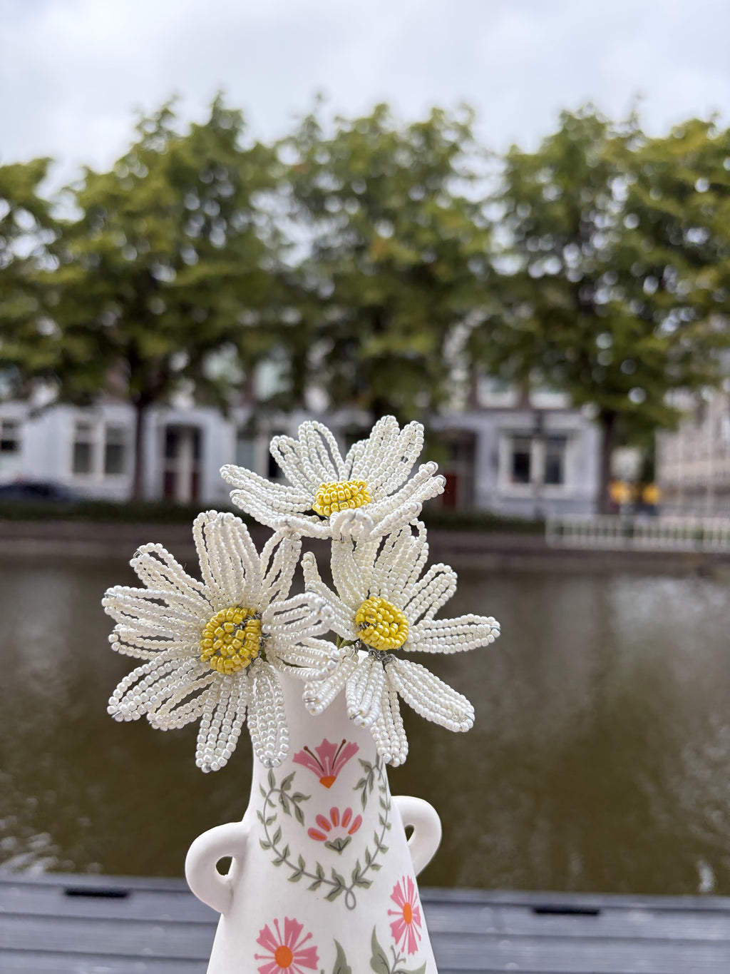 Three handmade beaded daisies with white petals and yellow centers in a hand-painted floral vase beside a canal