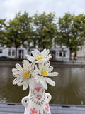 Three handmade beaded daisies with white petals and yellow centers in a hand-painted floral vase beside a canal